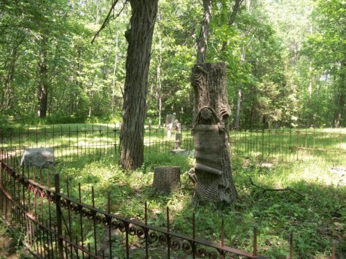 A serene cemetery scene with a tree stump, surrounded by a wrought iron fence and lush greenery.