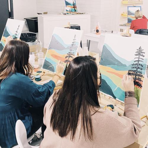 Two women painting landscapes on canvases in a bright, creative studio setting.