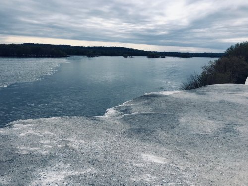 A calm river flows under a cloudy sky, with rocky banks and distant trees lining the horizon.