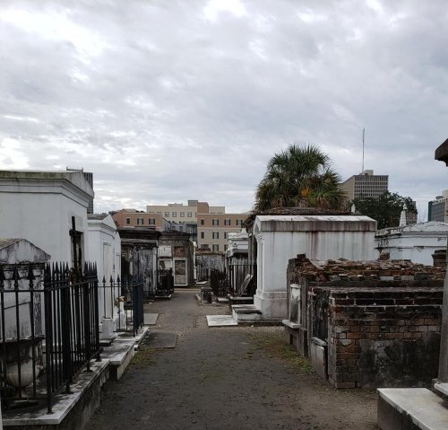A cemetery path lined with mausoleums and tombs, with a city skyline visible in the background under a cloudy sky.