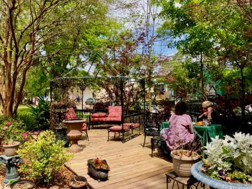 A serene garden patio with seating, surrounded by greenery and flowers, where two people enjoy a peaceful moment.