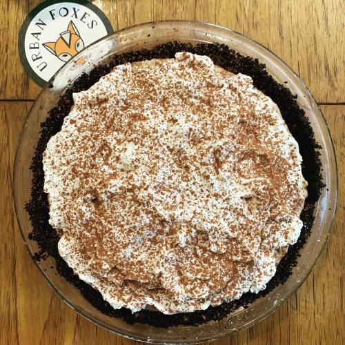 A chocolate pie topped with whipped cream and cocoa powder, placed on a wooden table next to a circular logo.