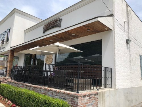 Exterior of a restaurant with a patio, featuring umbrellas and a sign that reads "District." Green shrubs in front.