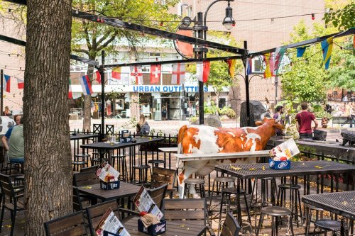 A lively outdoor dining area with tables, a cow statue, and colorful flags, surrounded by trees and shops.