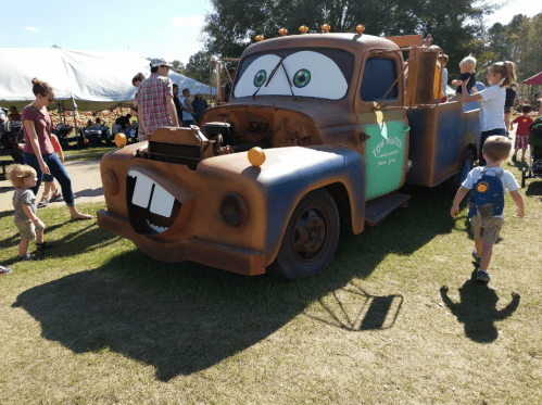 A colorful, cartoonish truck with a smiling face, surrounded by children and adults at an outdoor event.