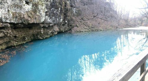 A serene blue river flows beside a rocky cliff, surrounded by bare trees and a peaceful natural landscape.