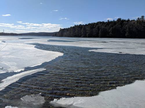 A serene winter scene of a partially frozen lake with rippling water and a forested shoreline under a clear blue sky.