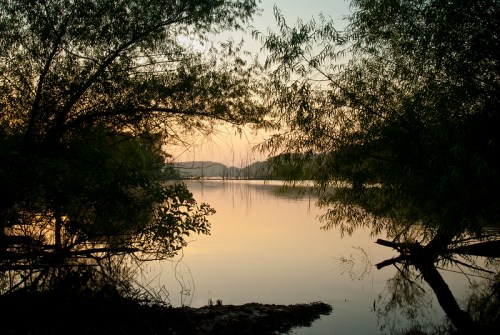 A serene lake at sunset, framed by trees and soft reflections on the water's surface.