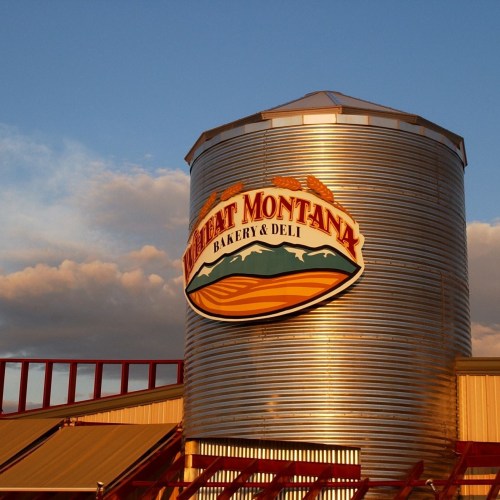 Sign for Wheat Montana Bakery & Deli on a silvery grain silo against a blue sky with clouds.