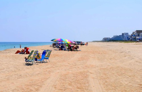 A sunny beach scene with colorful umbrellas, beach chairs, and people enjoying the sand and water.