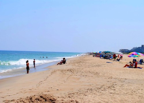 A sunny beach scene with people enjoying the water, lounging under umbrellas, and walking along the sandy shore.