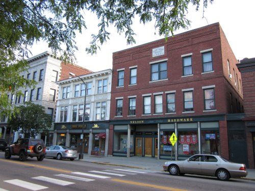 Historic brick buildings line a street, featuring shops like Nelson Hardware and a pool hall, with parked cars nearby.