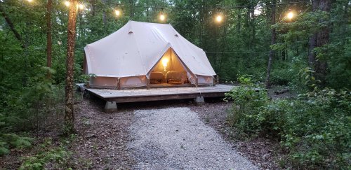 A glamping tent on a wooden platform, surrounded by trees and lit by string lights, with a warm glow from inside.