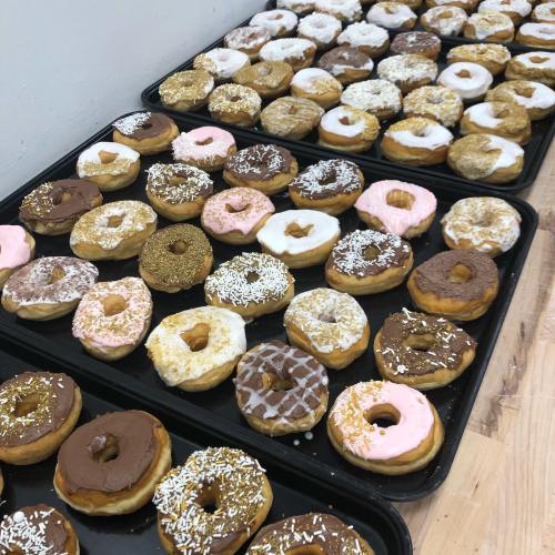 Trays of assorted donuts in various flavors and toppings, including chocolate, pink icing, and sprinkles.