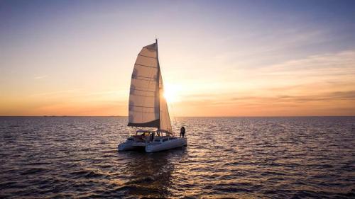 A sailboat with a large white sail glides across the water at sunset, casting a warm glow over the scene.