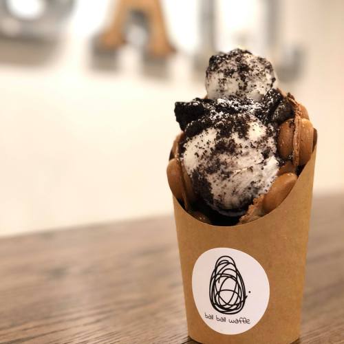 A waffle cone filled with two scoops of cookies and cream ice cream, topped with crushed Oreos, on a wooden table.