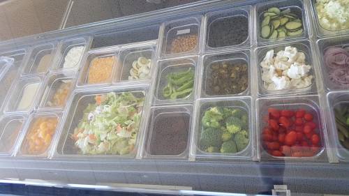 A variety of fresh salad ingredients in clear containers, including greens, tomatoes, cucumbers, and broccoli.