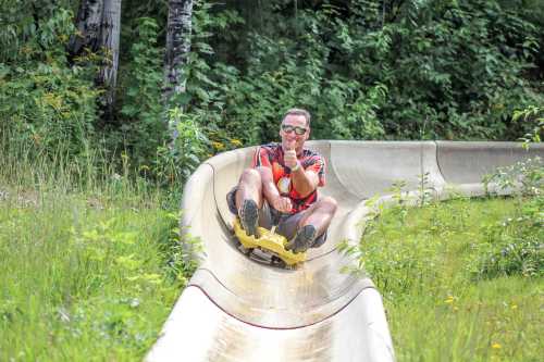 A man in sunglasses slides down a water slide surrounded by greenery, smiling and giving a thumbs-up.