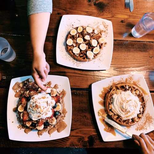 Three plates of waffles topped with bananas, berries, whipped cream, and drizzled with syrup on a wooden table.