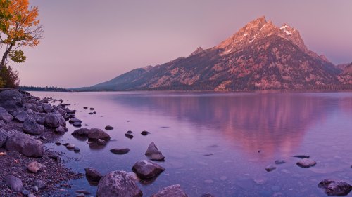 A serene lake reflects a mountain at sunset, surrounded by rocky shores and autumn-colored trees.