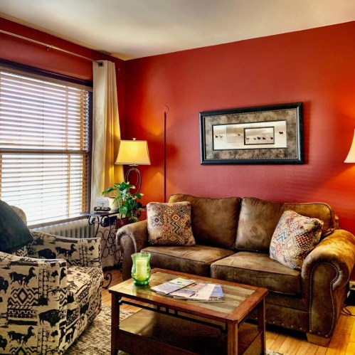 Cozy living room with a brown couch, patterned chair, wooden coffee table, and warm red walls. Natural light from windows.