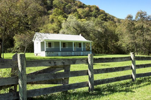 A quaint house with a porch sits beside a wooden fence, surrounded by lush greenery and rolling hills.