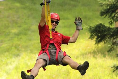 A person in a red shirt and helmet zip-lining through a green landscape, smiling and waving.