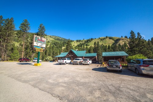 A rustic lodge surrounded by trees and mountains, with parked cars and a clear blue sky.