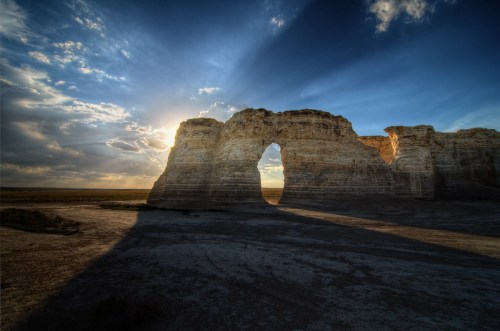 A dramatic rock formation with an arch, illuminated by sunlight against a blue sky with scattered clouds.