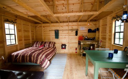 Cozy wooden cabin interior with a bed, dining table, and kitchenette, featuring natural light from windows.