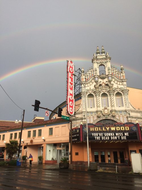 A historic Hollywood theater under a double rainbow, with a cloudy sky and street signs nearby.
