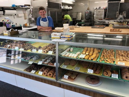 A smiling young boy stands behind a glass display filled with various baked goods, including donuts and pastries.