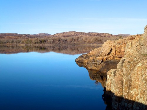 A calm lake reflects rocky shores and distant hills under a clear blue sky.