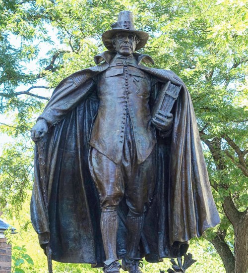 A bronze statue of a man in a hat and cape, holding a book, surrounded by trees.