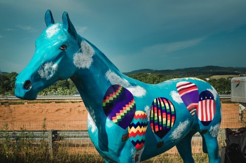 A blue horse statue decorated with colorful hot air balloon designs against a scenic landscape.