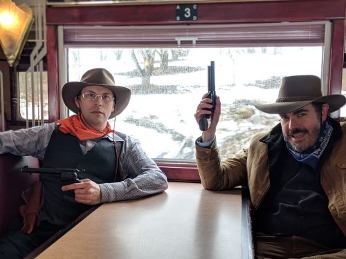 Two men in cowboy attire sit in a train car, each holding a gun, with a snowy landscape visible through the window.