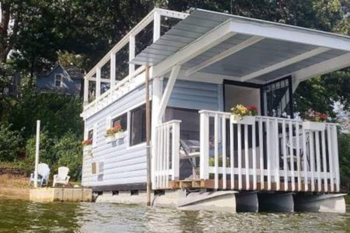 A small blue houseboat with a deck, surrounded by water and greenery, featuring flower boxes and outdoor seating.