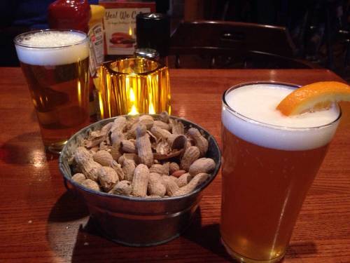Two glasses of beer sit on a wooden table next to a bowl of peanuts and a lit candle.