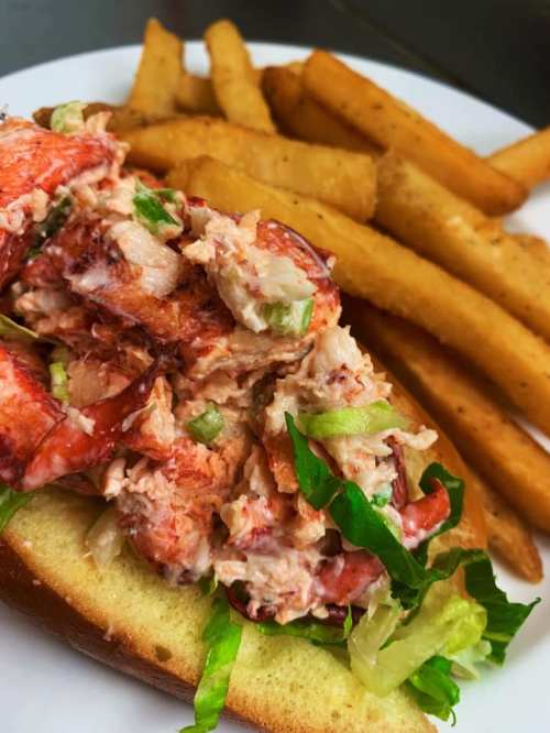 A lobster roll topped with lettuce, served with golden-brown fries on a white plate.