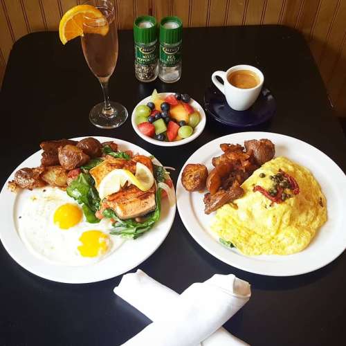 Two plates of breakfast: one with salmon, greens, and eggs; the other with an omelet, potatoes, and fruit. A drink and coffee are nearby.