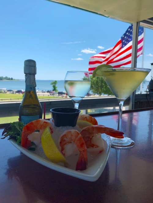 A plate of shrimp on ice with lemon, two cocktails, and an American flag in the background by a scenic waterfront.