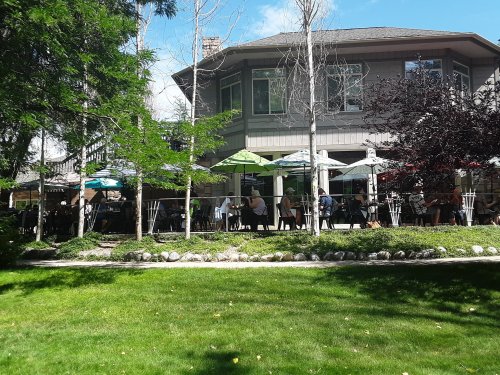 A lively outdoor dining area with umbrellas, surrounded by greenery and a large building in the background.