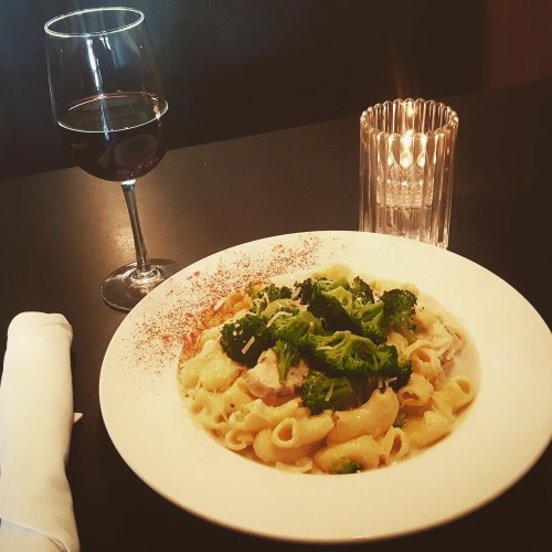 A plate of creamy pasta with broccoli, accompanied by a glass of red wine and a candle on a dark table.