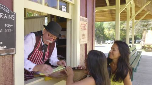 A man in a bowler hat assists two women at a ticket window in a rustic setting.