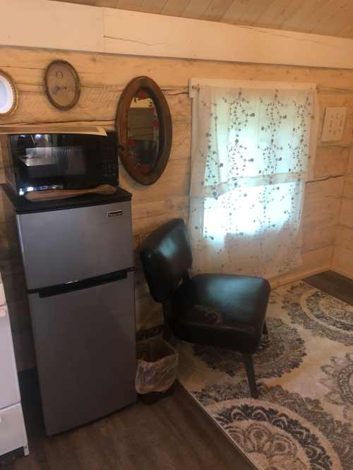 A cozy corner of a room featuring a small fridge, microwave, a chair, and a window with sheer curtains.