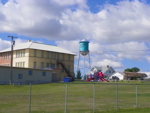 A water tower stands behind a school building, with a playground and houses in the background under a cloudy sky.