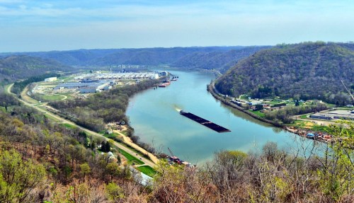 Aerial view of a river winding through green hills, with industrial buildings and a barge navigating the water.
