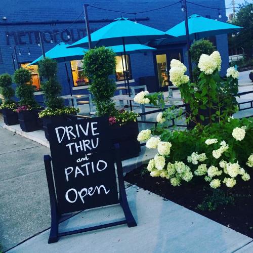 A sign reads "Drive Thru and Patio Open" outside a restaurant with blue umbrellas and blooming white flowers.