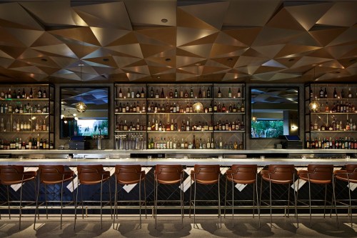 A modern bar with a geometric ceiling, lined shelves of liquor, and stylish brown bar stools.