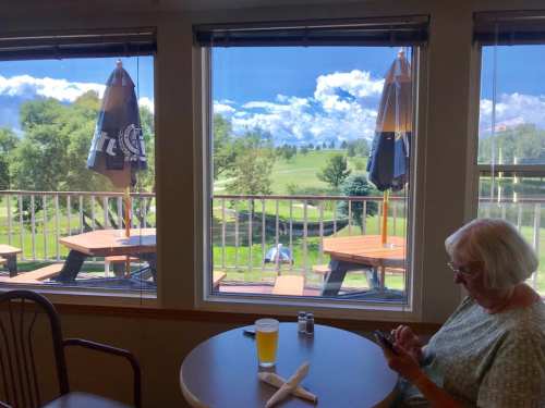 A woman sits at a table with a drink, looking at her phone, with a scenic view of green hills and blue skies outside.